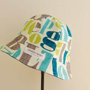 May include: A white bucket hat with a colorful alphabet print in shades of blue, green, and gray. The hat has a brown label that reads "Dakko Baby".