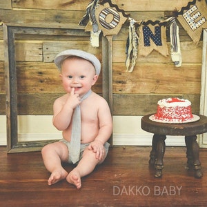 May include: A baby boy wearing a gray and white striped tie and a gray hat sits on a wooden floor in front of a rustic wooden wall. A red and white frosted cake sits on a small wooden stool. The wall is decorated with a banner that says "ONE" in blue and white letters.