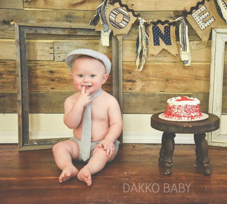 May include: A baby boy wearing a gray and white striped tie and a gray hat sits on a wooden floor in front of a rustic wooden wall. A red and white frosted cake sits on a small wooden stool. The wall is decorated with a banner that says "ONE" in blue and white letters.