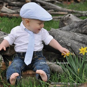 May include: A young child wearing a blue and white striped hat, a white button-down shirt with a blue and white striped tie, and blue jeans is sitting on a wooden log and reaching for a yellow daffodil.