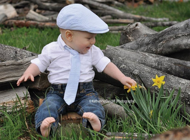 May include: A young child wearing a blue and white striped hat, a white button-down shirt with a blue and white striped tie, and blue jeans is sitting on a wooden log and reaching for a yellow daffodil.