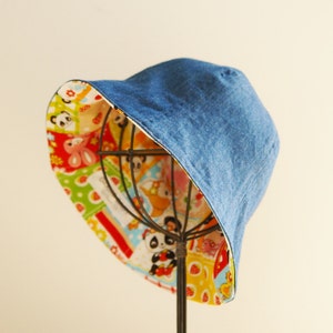 Puede incluir: Un sombrero de cubo de mezclilla azul con un forro de estampado de dibujos animados coloridos. El sombrero est&aacute; en un soporte.
