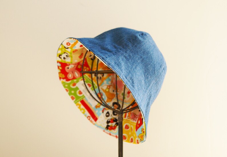 Puede incluir: Un sombrero de cubo de mezclilla azul con un forro de estampado de dibujos animados coloridos. El sombrero est&aacute; en un soporte.