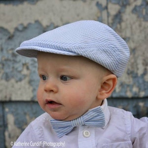 May include: A baby boy wearing a white button-down shirt with a blue and white striped bow tie and a blue and white striped hat. The baby is looking to the left of the camera.