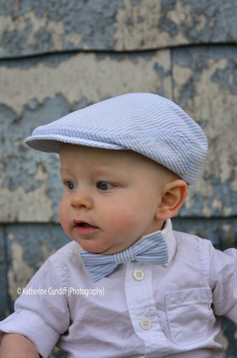 May include: A baby boy wearing a white button-down shirt with a blue and white striped bow tie and a blue and white striped hat. The baby is looking to the left of the camera.