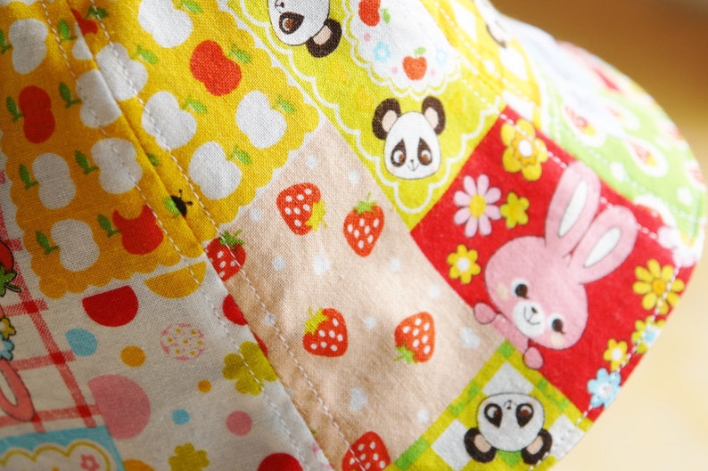 Puede incluir: Un sombrero de tela colorido con un dise&ntilde;o de patchwork que presenta fresas, flores y osos panda. El sombrero est&aacute; hecho de algod&oacute;n y tiene una combinaci&oacute;n de colores rojo, amarillo y rosa.