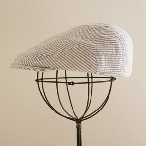 May include: A white and gray striped cotton newsboy cap with a brown brim. The cap is displayed on a black metal stand.