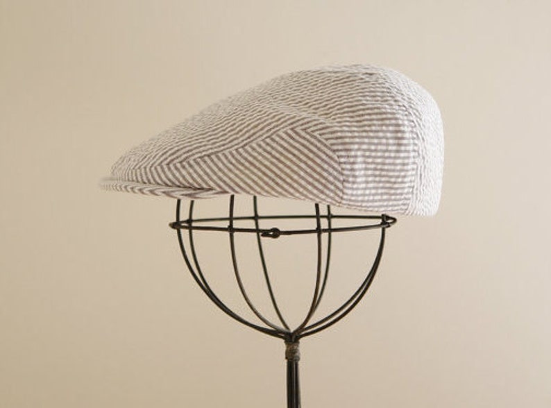 May include: A white and gray striped cotton newsboy cap with a brown brim. The cap is displayed on a black metal stand.