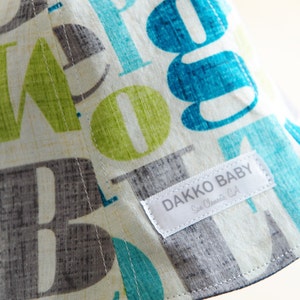 May include: A white sun hat with a blue, green, and gray alphabet print. The hat has a label that reads "Dakko Baby, San Clemente, CA".