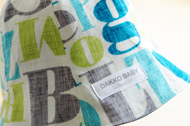 May include: A white sun hat with a blue, green, and gray alphabet print. The hat has a label that reads "Dakko Baby, San Clemente, CA".