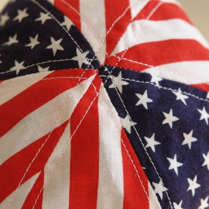 May include: Close-up of the top of a red, white, and blue fabric hat with a star pattern. The hat is made of a soft, cotton fabric and has a sewn seam in the center.