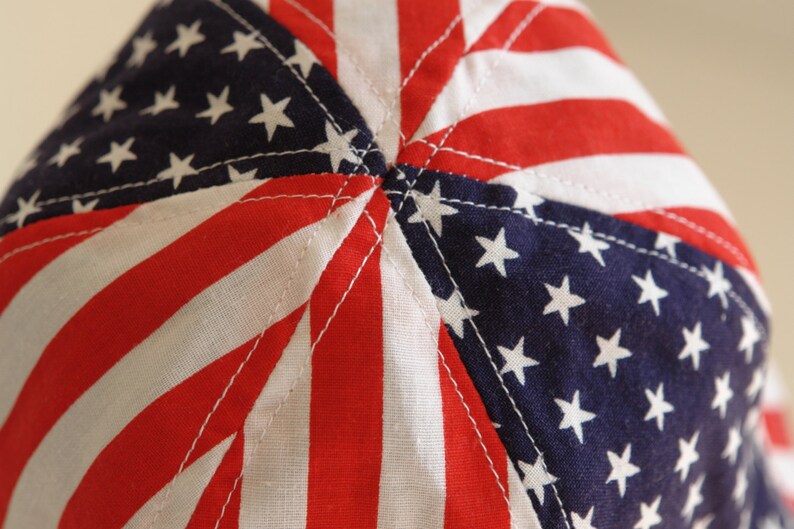 May include: Close-up of the top of a red, white, and blue fabric hat with a star pattern. The hat is made of a soft, cotton fabric and has a sewn seam in the center.