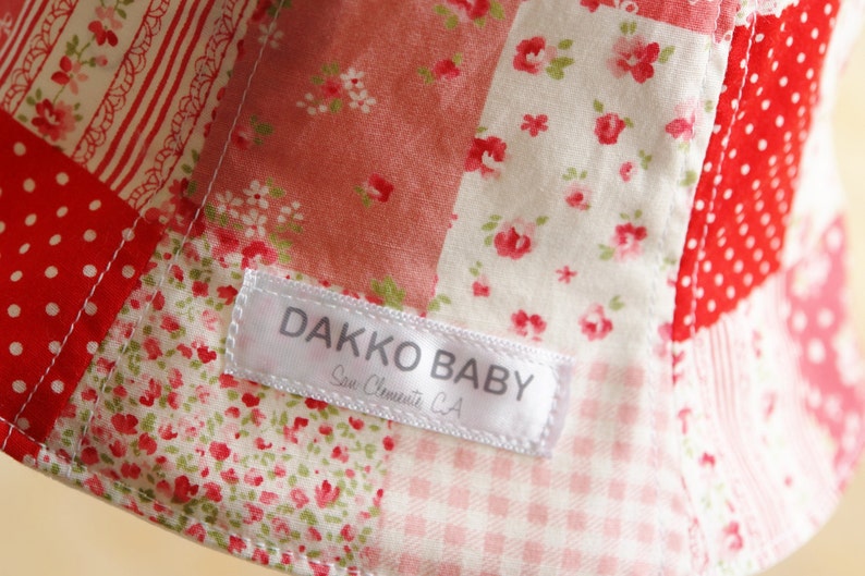 May include: A white label with the text "DAKKO BABY" and "San Clemente, CA" sewn onto a red and white patchwork fabric with floral patterns.