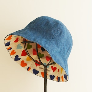 Bucket-styled denim hat is displayed in front of the ivory color wall.  It shows the inside of the hat, a colorful heart pattern on it.