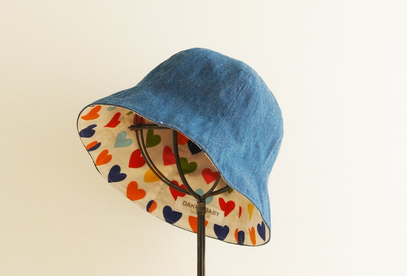Bucket-styled denim hat is displayed in front of the ivory color wall.  It shows the inside of the hat, a colorful heart pattern on it.