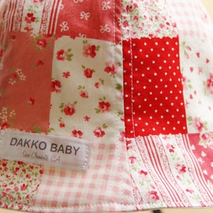May include: A red, white, and pink patchwork baby hat with a white label that reads "Dakko Baby, San Clemente, CA".