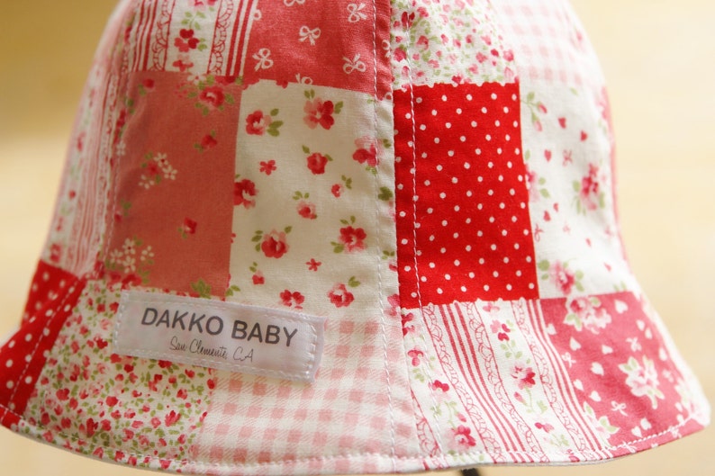 May include: A red, white, and pink patchwork baby hat with a white label that reads "Dakko Baby, San Clemente, CA".