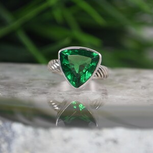 May include: A silver ring featuring a large, triangular-cut, vibrant green gemstone set in a polished metal frame. The band of the ring displays a distinct twisted, cable-like texture that wraps around the finger, providing a detailed metallic finish.