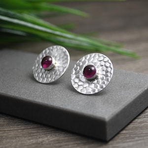 May include: A pair of silver earrings with a hammered texture and a round, dark purple gemstone in the center of each earring.