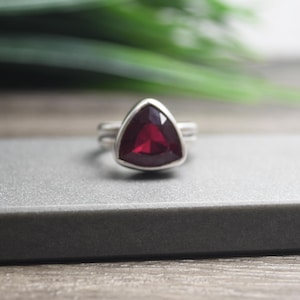 Op de afbeelding: Een zilveren ring met een driehoekige rode edelsteen.