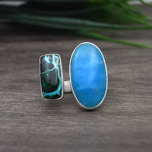 May include: A silver ring with two gemstones. The larger stone is oval shaped and a light blue color. The smaller stone is rectangular and a dark green color with white veins.