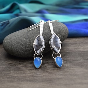 May include: A pair of silver dangle earrings featuring a marquise-shaped dendritic opal stone with black fern-like inclusions, topped by a textured silver bar and finished with a small, smooth, teardrop-shaped blue chalcedony stone at the base.