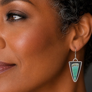 May include: A triangular drop earring featuring a polished silver frame with a dark oxidized border. The center inlay is a smooth, variegated stone displaying horizontal bands of teal, green, and cream colors, suspended from a simple silver hook.