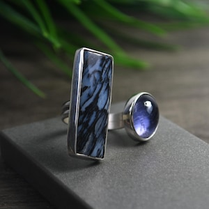 May include: A silver ring featuring two distinct stones: a tall, rectangular stone with a black and white marbled pattern and a smaller, round, translucent purple stone. The metal band is open, connecting both stone settings in a bypass design.