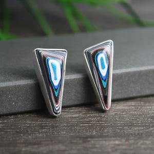 May include: A pair of triangular stud earrings featuring layered, multi-colored patterns of black, white, red, and metallic blue. The earrings are set in a silver-toned metal frame, showcasing a polished, smooth surface with concentric, abstract designs.