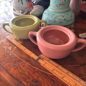 May include: Two vintage ceramic sugar bowls with lids. The bowl on the left is a bright green color and the bowl on the right is a pale pink color. Both bowls have a simple, rounded shape with two small handles.