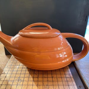 May include: A bright orange ceramic teapot with a rounded, slightly curved body and a lid. The teapot has a handle on the side and a spout on the front.
