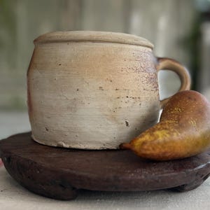 May include: A rustic, beige ceramic mug with a lid and handle, resting on a dark wooden plate. A ripe pear with yellow and brown hues sits beside the mug. The mug and pear are set against a blurred background.