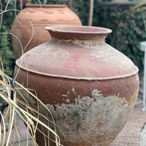 May include: Two large, weathered terracotta pots with a rustic, aged appearance. The foreground pot is a reddish-brown with visible texture and a wide opening. The background pot is a lighter brown. The pots are outdoors, surrounded by greenery.