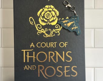 May include: A black hardcover book with gold lettering. The title is "A Court of Thorns and Roses" by Sarah J. Maas. A gold rose with thorns is on the cover. A small, dark green charm with a gold outline is attached to the book.
