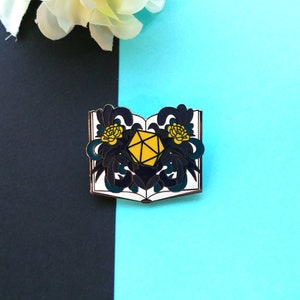 May include: An enamel pin featuring a book with a black and white floral design and a yellow d20 die in the center.