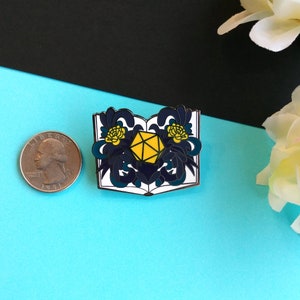 May include: A white enamel pin with a blue and yellow design. The pin features an open book with a yellow 20-sided die in the center. The book is surrounded by blue and yellow floral designs.