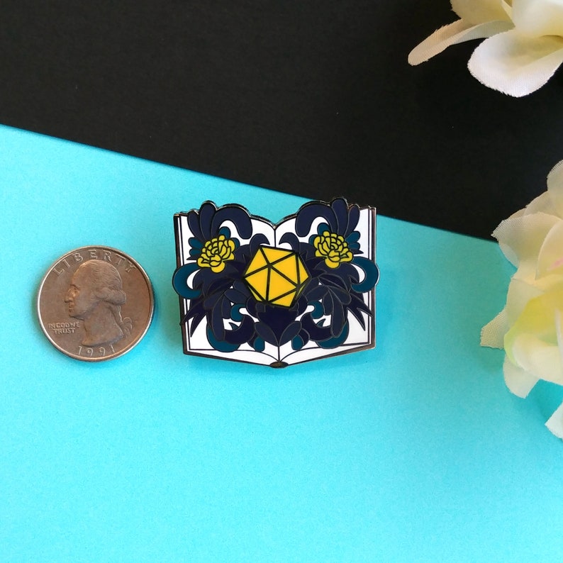 May include: A white enamel pin with a blue and yellow design. The pin features an open book with a yellow 20-sided die in the center. The book is surrounded by blue and yellow floral designs.