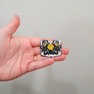 May include: A silver enamel pin featuring a book with a yellow and black d20 die inside. The book is open and has black floral designs on the pages.