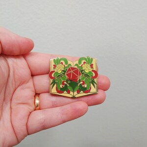 May include: Gold-tone enamel pin featuring a red twenty-sided die surrounded by green leaves and yellow flowers, all within an open book.