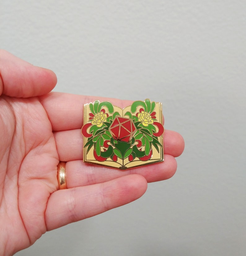 May include: Gold-tone enamel pin featuring a red twenty-sided die surrounded by green leaves and yellow flowers, all within an open book.