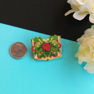 May include: Gold-tone enamel pin featuring a red twenty-sided die inside an open book with green and yellow floral details.
