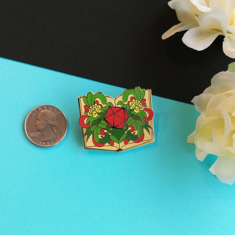 May include: Gold-tone enamel pin featuring a red twenty-sided die inside an open book with green and yellow floral details.