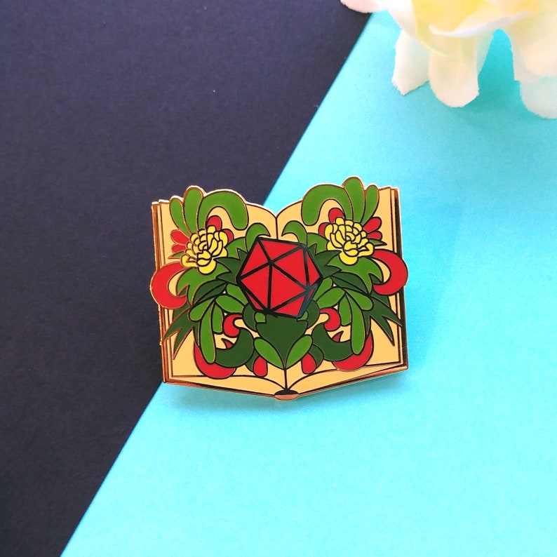 May include: Gold-tone enamel pin of an open book with a red and black dice in the center. The book is surrounded by green leaves and yellow flowers.