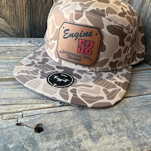 May include: A five-panel baseball cap with a tan and brown camouflage pattern. A rectangular brown leather patch is centered on the front, featuring the black text Engine 52 and Jacksonville Fire Division, with the number 52 in red. A black sticker is on the brim.
