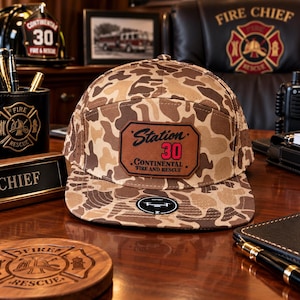 May include: A five-panel baseball cap featuring a brown and tan camouflage pattern. A rectangular leather patch on the front displays the text Station 30 Continental Fire and Rescue in black and red lettering. A circular sticker is on the flat brim.