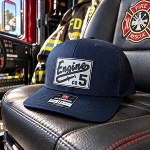 May include: A navy blue trucker hat featuring a rectangular patch with the text Engine Co 5 and a graphic of a fire hose nozzle. The hat has a structured front panel, a mesh back, and a black sticker on the brim that reads Richardson 112.