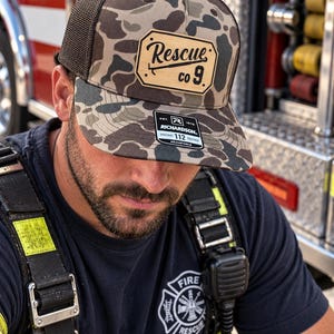 May include: A camouflage trucker hat with a brown mesh back and a tan leather-like patch on the front. The patch features the text Rescue co 9 with a graphic of a tool. A black sticker on the brim displays the text Richardson 112 Adjustable.