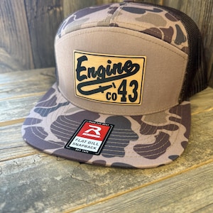 May include: A flat bill snapback hat with a brown and camouflage pattern. The hat features a brown mesh back and a rectangular patch with the words "Engine co 43" in black. A black and white tag is attached to the brim.