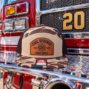 May include: A camouflage print snapback hat with a brown mesh back and a tan front panel. A leather patch on the front features the text COOL SPRINGS EST 1960 FIRE DEPT. and a central emblem with the number 20. A small rectangular sticker is on the brim.