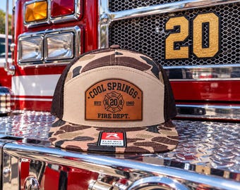 May include: A camouflage print snapback hat with a brown mesh back and a tan front panel. A leather patch on the front features the text COOL SPRINGS EST 1960 FIRE DEPT. and a central emblem with the number 20. A small rectangular sticker is on the brim.